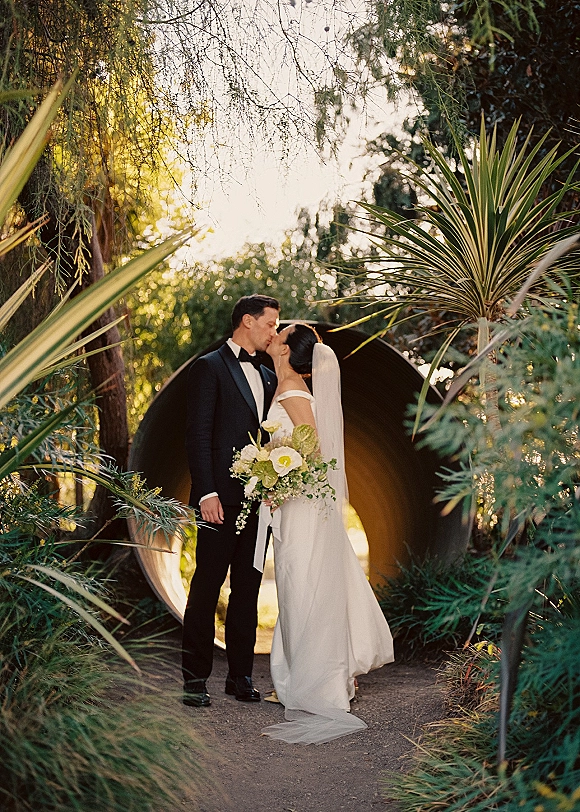 Wedding kiss portrait of bride and groom kissing under her veil, holding a calla lily bouquet on a garden path with a circular tunnel backdrop