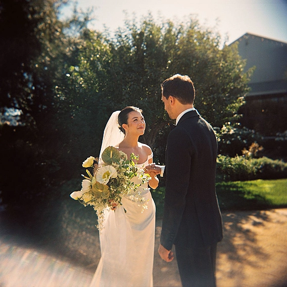 Couple portrait of bride and groom holding hands, bride in strapless dress and veil with bouquet, backlit in garden greenery at sunset