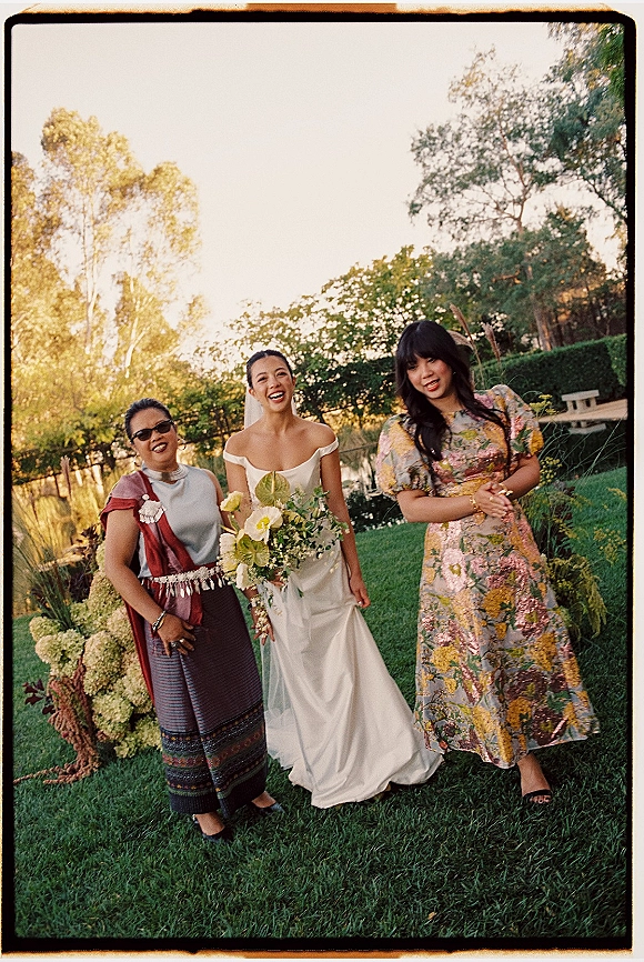 Bride with guests smiling together, holding a white and green bouquet, in a garden lawn setting with trees and floral arrangements