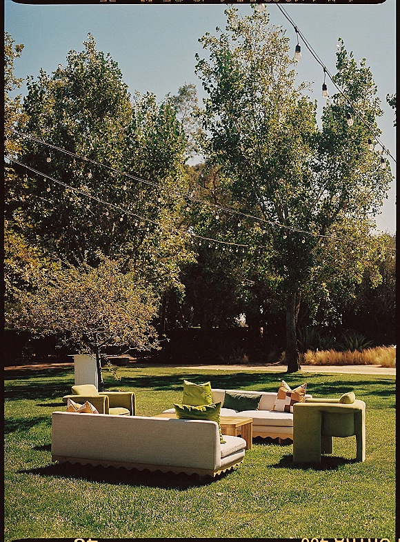 Lounge seating area with wedding lounge seating sofas, chairs, and colorful throw pillows around a low table under string lights on lawn
