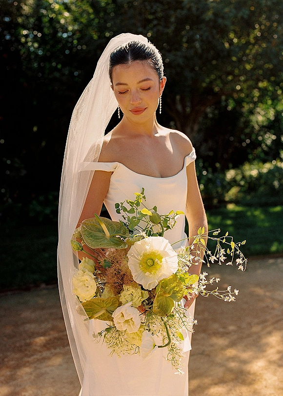 Bridal portrait of a bride holding bouquet in an off-the-shoulder satin dress with veil, drop earrings, and sunlit garden greenery backdrop