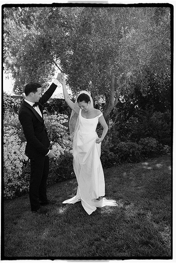 Couple portrait in a black and white wedding photo, groom in tuxedo spinning bride in veil on a garden lawn among trees