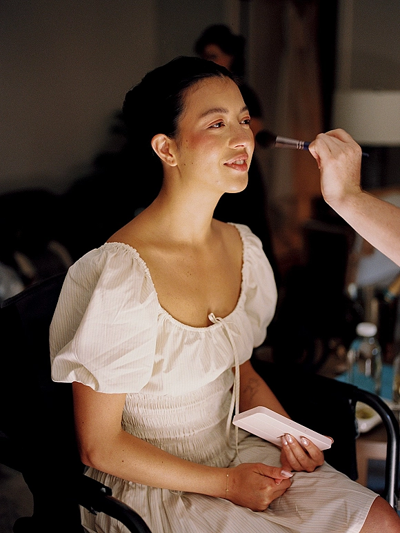 Bridal makeup in progress as a wedding makeup artist applies with a brush, bride in robe with updo seated at vanity doorway backdrop