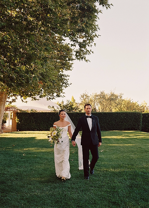 Couple portrait of bride and groom walking hand in hand across a garden lawn, bride holding bouquet with long veil, groom in tuxedo