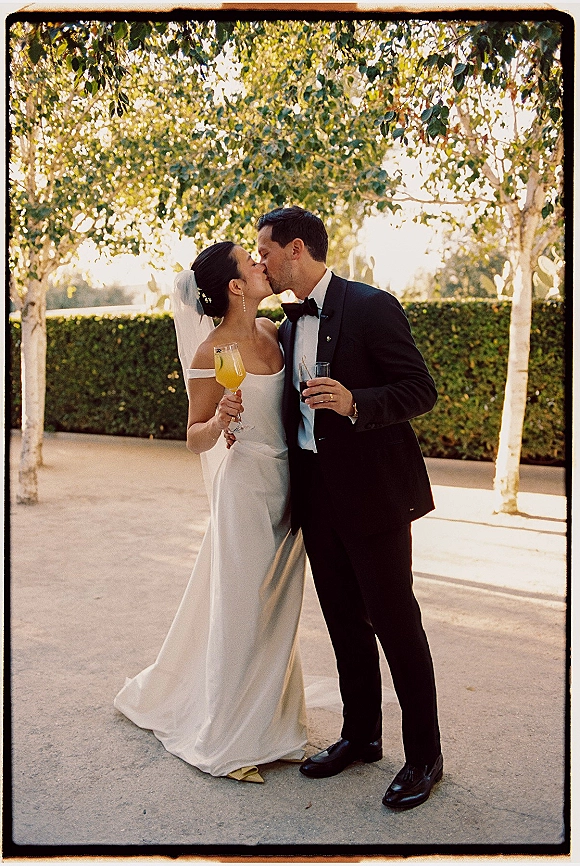 Wedding kiss portrait of bride and groom kissing, holding champagne flute and cocktail glass on a patio with trees and green hedge backdrop