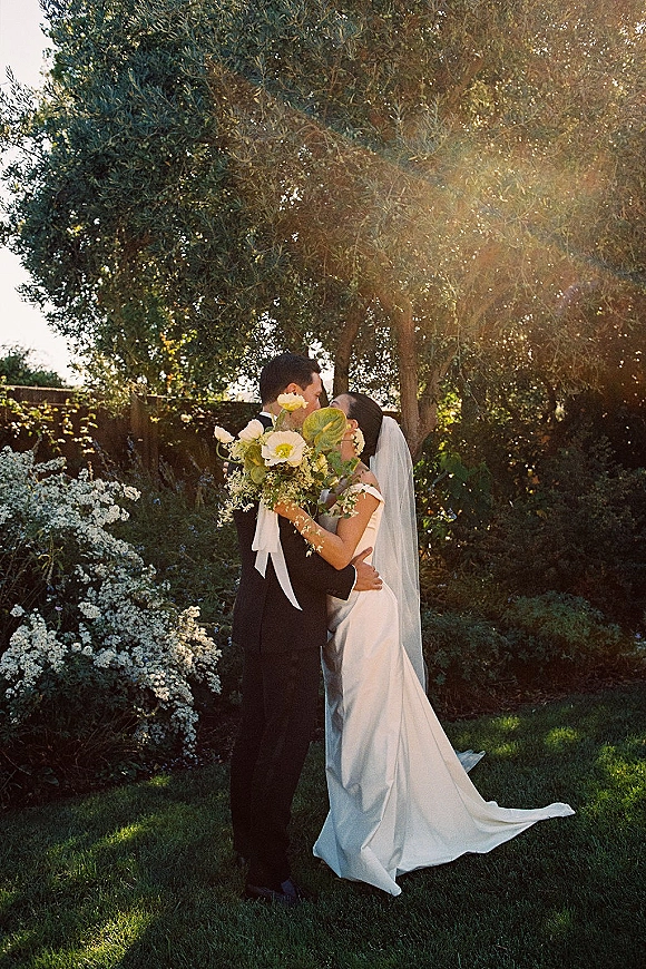 Wedding kiss portrait of bride and groom kissing behind a bouquet, her long veil flowing as sun flare lights a garden lawn with shrubs and trees