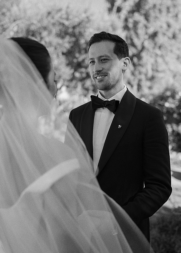 Wedding vows as groom in tuxedo with bow tie and boutonniere looks at bride through her veil, framed by garden trees