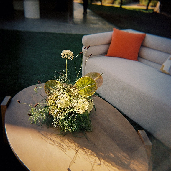 Cocktail lounge decor with a curved outdoor sofa and orange throw pillow around a round coffee table on a grassy patio with florals