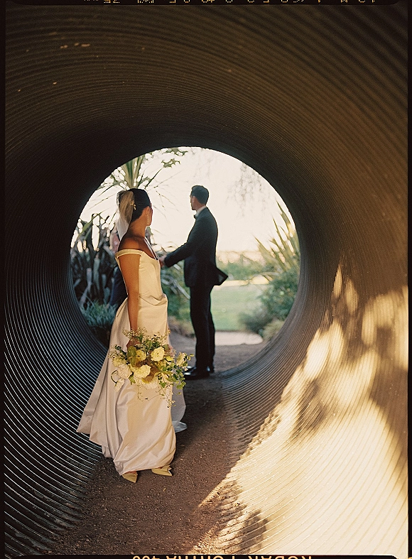 Wedding first look as bride in off-the-shoulder satin dress and veil approaches groom in black suit along a sunlit tunnel path