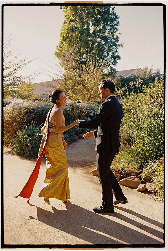 Couple portrait of bride and groom holding hands on a garden path, bride in a gold dress with scarf shawl and hair bun, groom in tuxedo