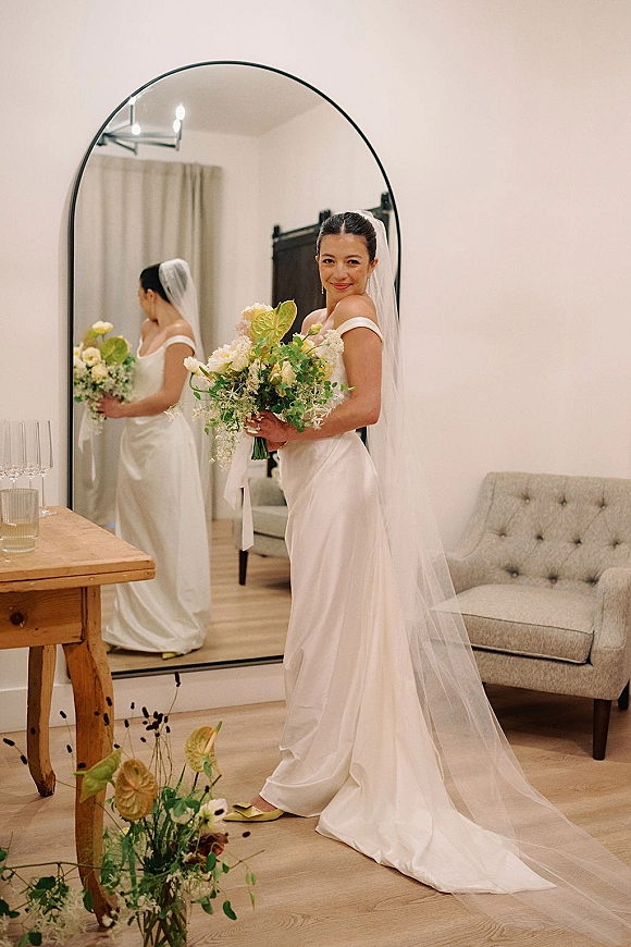 Bridal portrait of a bride holding bouquet in a strapless wedding dress with long veil, standing by an arched mirror in a bright bedroom