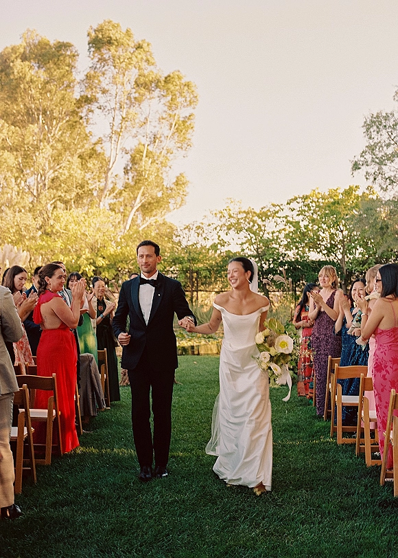 Wedding recessional as bride and groom walk the grass aisle hand in hand, bride in off the shoulder dress with veil and bouquet, guests clapping in garden setting