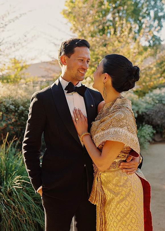 Couple portrait of bride and groom embrace, he in black tuxedo and bow tie, she in embellished gold sari, in sunlit garden