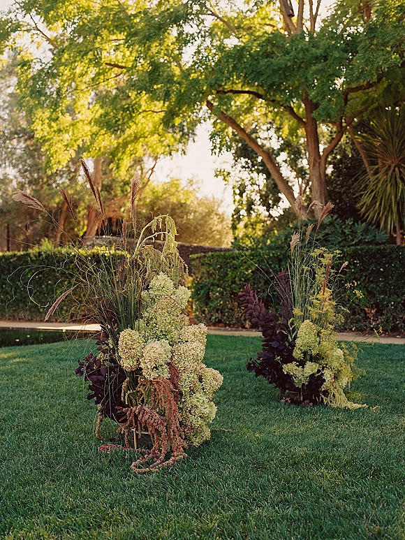 Ceremony aisle florals with ground floral aisle markers of hydrangeas, pampas grass, greenery, and burgundy foliage on a sunny lawn path