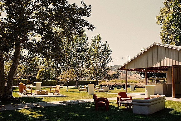 Outdoor lounge setup with sofas and lounge chairs around coffee tables on a grass lawn, with string lights and hills beyond the pavilion