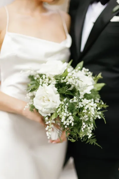 Wedding bouquet of white roses and lily of the valley with greenery, held by bride in satin straps beside groom in black tuxedo