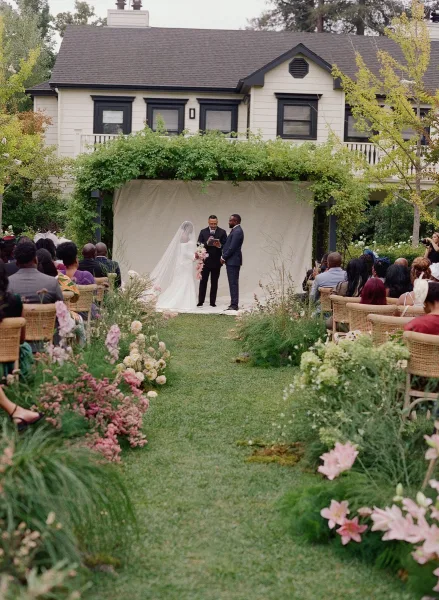 Wedding ceremony with bride and groom under a fabric-draped arch with greenery accents, aisle flowers and guests on a garden lawn