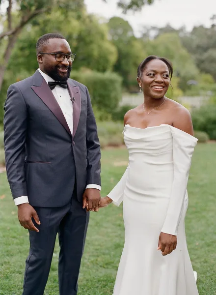 Couple portrait in a garden, bride and groom holding hands, her wedding dress and his tuxedo with glasses amid lush greenery