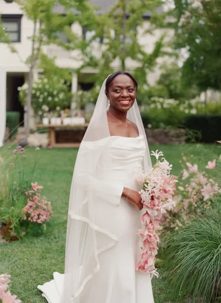 Bridal portrait of a bride holding bouquet with a cathedral veil, smiling on a garden lawn with greenery and flowering plants behind her