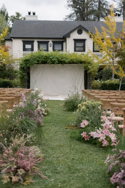 Outdoor ceremony setup for a garden wedding ceremony with wicker chairs facing a draped fabric backdrop, greenery garland, and ground florals on a lawn aisle runner