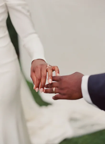 Wedding rings in a ring exchange moment, engagement ring and band on groom’s hand with suit jacket and tie against bright lawn sky