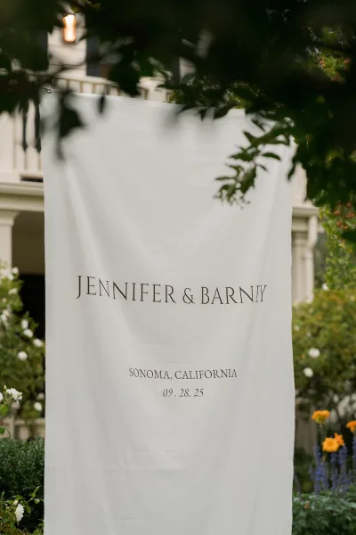 Wedding welcome sign on a fabric banner with black lettering, hanging at a garden entrance near porch railing and flowering greenery