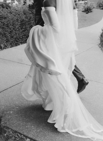 Recessional moment as bride and groom walking away on a sidewalk, her long veil and dress train flowing past hedges and lawn