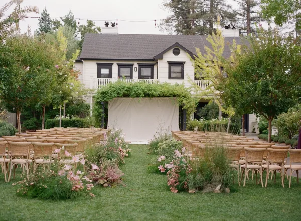Outdoor ceremony setup with garden wedding ceremony seating, wicker chairs lining a floral aisle toward a fabric backdrop with string lights on a lawn