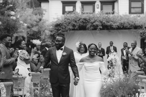 Wedding recessional as bride and groom walking aisle hand in hand, bouquet and veil flowing as guests raise phones at an outdoor garden ceremony