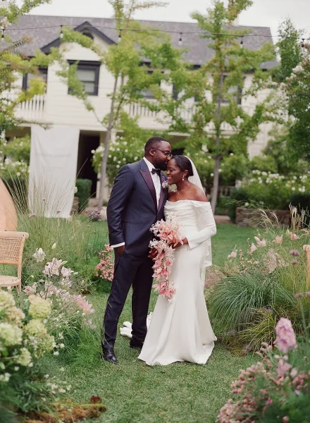Couple portrait of groom kissing bride’s forehead as they hold a cascading bouquet, her veil draping in a garden by a white facade