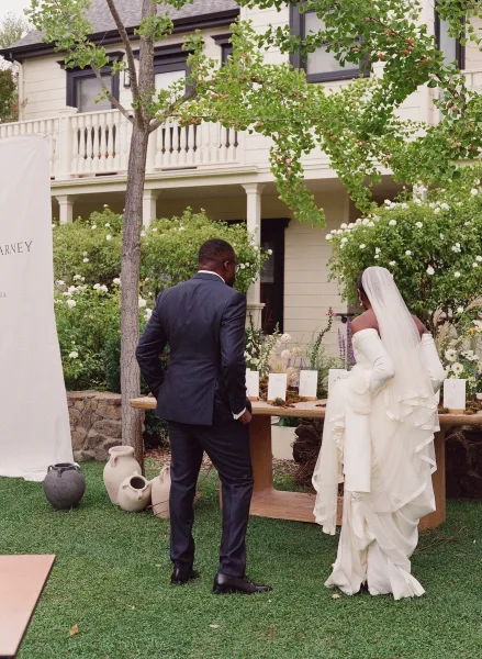 Wedding escort display and escort card table with textured white cards, ceramic vases and potted plants as bride and groom view in a garden lawn setting