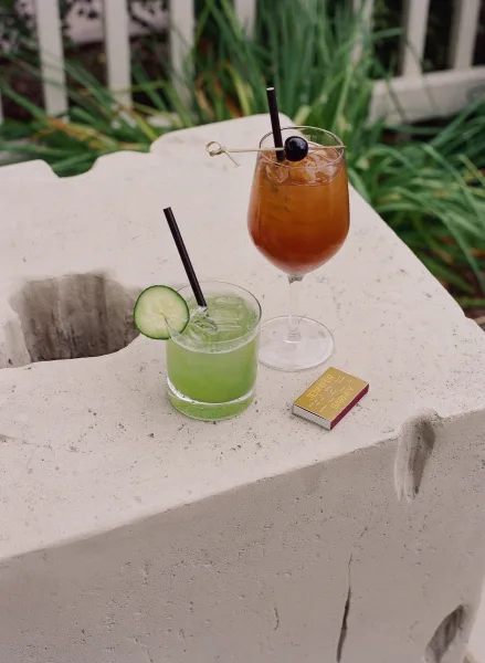 Wedding cocktails with signature wedding cocktails in coupe and rocks glasses, black straws, cucumber and cherry garnish on stone by greenery
