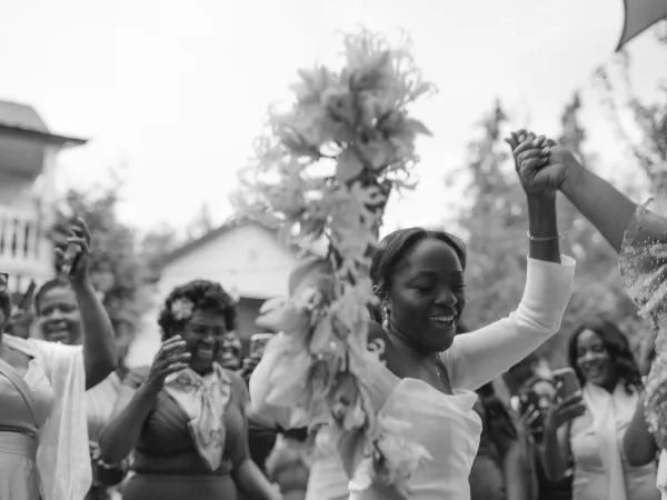 Wedding celebration moment as bridesmaids cheering with raised hands, captured among guests holding phones outdoors by trees and a house, bracelet accent