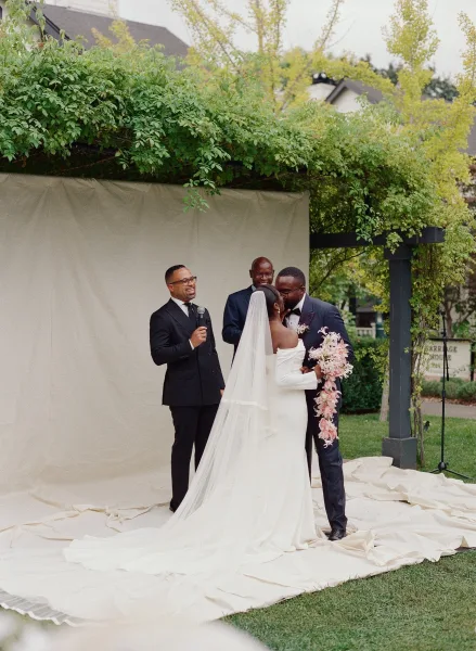 Ceremony kiss as bride and groom embrace beneath a greenery-covered pergola, her long veil and blush bouquet trailing by a draped backdrop