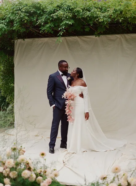 Couple portrait of bride in off the shoulder wedding dress and groom in black tuxedo holding a cascading bouquet before beige fabric backdrop outdoors