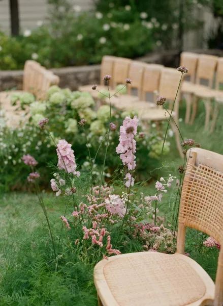 Ceremony aisle florals with meadow aisle flowers in pink, white, and greenery line wood chairs with cane backs on a garden lawn near a stone wall