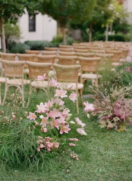 Ceremony aisle decor with woven chair rows and low pink flower clusters, set on a garden lawn with trees and a white building backdrop