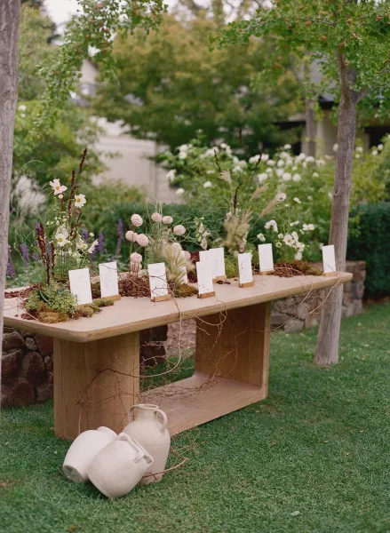 Escort card display with wedding seating chart cards on a wood table, moss and wildflowers in ceramic jugs, set in a garden yard by a stone wall
