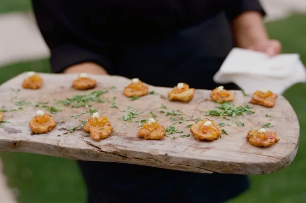 Wedding passed appetizers with chutney and herb garnish on a wood serving board, carried by catering staff on an outdoor lawn with greenery