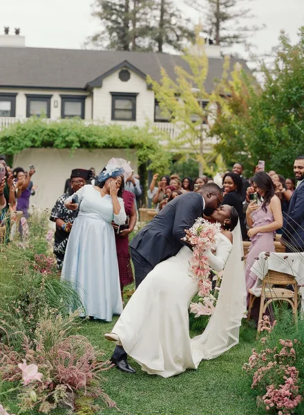 Wedding kiss portrait of bride and groom in a dip kiss wedding photo, her long veil and cascading bouquet as guests cheer on a garden lawn