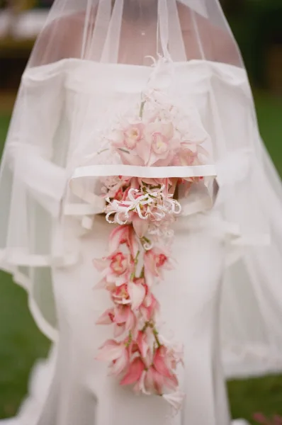 Wedding bouquet in a cascading wedding bouquet style with soft pink flowers and white ribbon, held against a veil and green lawn background