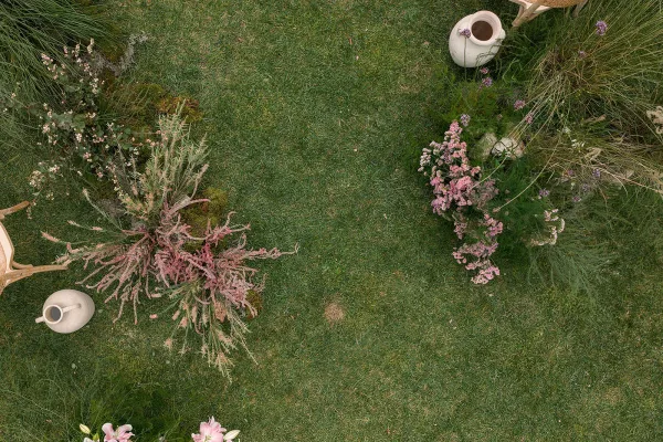 Ceremony aisle design with garden ceremony aisle ground florals in pink blooms and greenery, ceramic vases beside woven chairs on a grass lawn