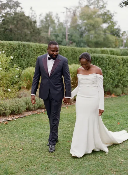 Couple portrait of bride and groom walking hand in hand on a garden lawn, her off-the-shoulder gown with train beside his tuxedo bow tie