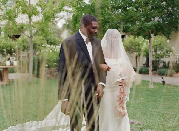 Wedding couple portrait of bride and groom walking on a garden lawn, her long veil over her face and cascading bouquet, his black tuxedo and bow tie