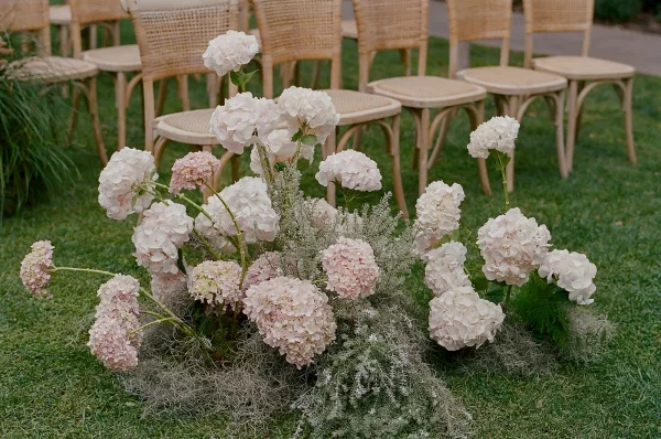 Ceremony aisle florals and ground floral arrangement with white and blush hydrangeas and ferns lining a grass lawn walkway with rattan chairs