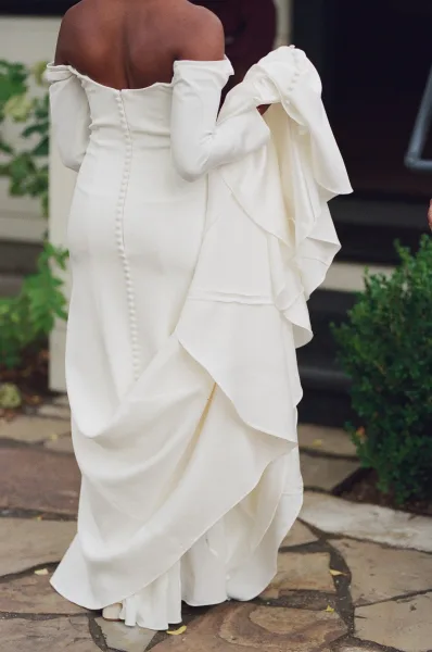 Wedding dress with off the shoulder sleeves and button-back detail, shown from behind with a flowing train on a stone walkway by greenery