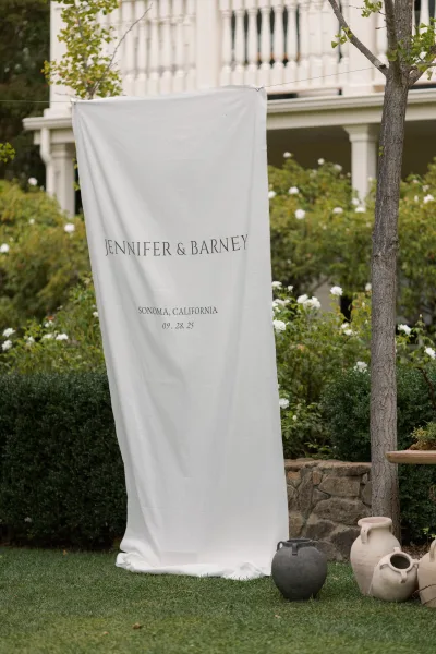 Wedding welcome sign with a fabric wedding welcome sign banner, black lettering on hanging linen with rope line beside ceramic urns in a garden