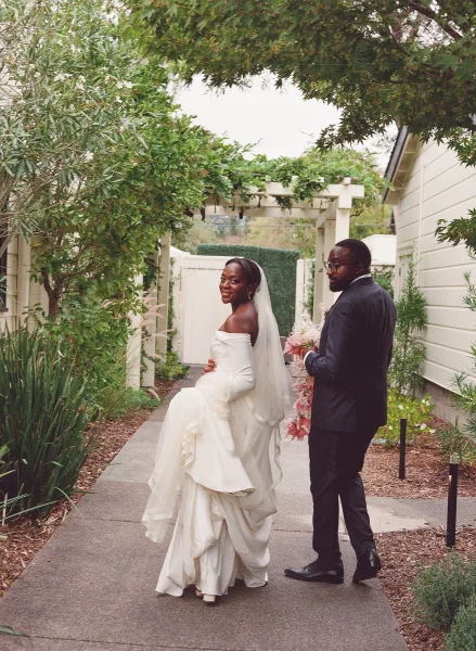 Couple portrait of bride in off-the-shoulder gown and long veil with groom in black suit walking a pergola garden walkway, bouquet in hand