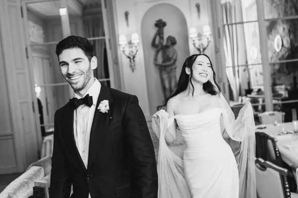 Couple portrait in a black and white wedding photo, the bride in a fitted gown with sheer sleeves beside her groom, against mirrors and sconces.