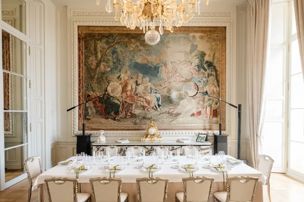 Reception tablescape on a long banquet table with white and gold place settings and candles, set before a tapestry wall and tall windows.